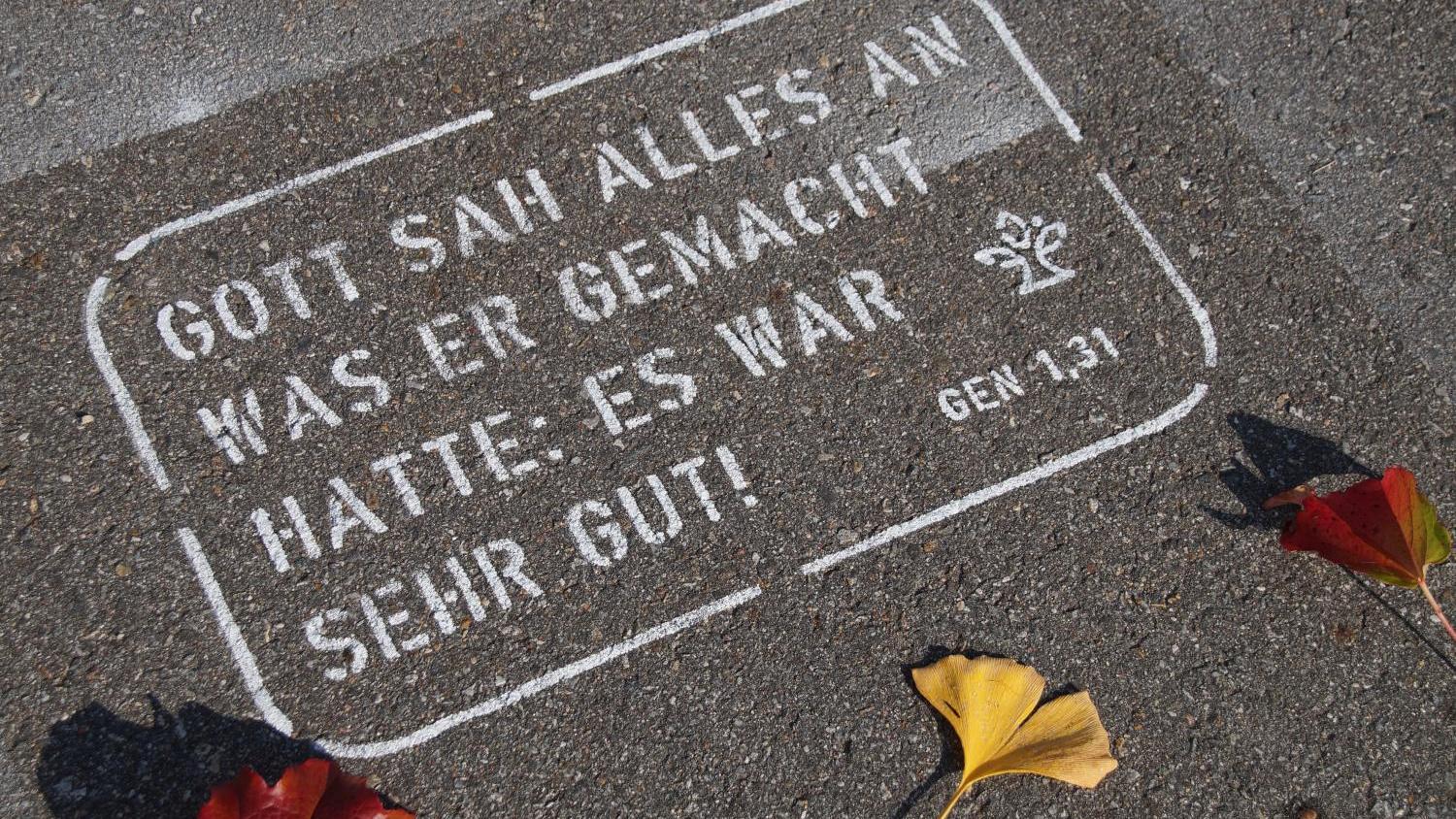 bibel-graffiti_02_1_by_rudi_berzl_pfarrbriefservice Weißer Bibelvers auf Asphalt, umgeben von bunten Herbstblättern.
