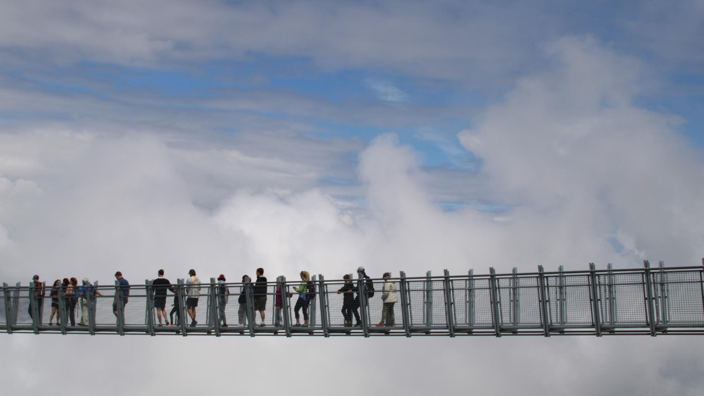 Menschen gehen über eine Hängebrücke. Im Hintergrund sind Wolken zu sehen